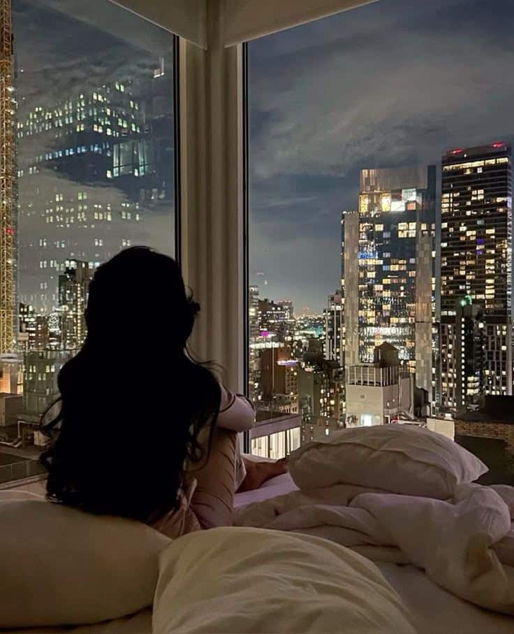 Cant sleep - Handy Home Woman sitting on a bed, gazing at a cityscape through a large window at night, with illuminated skyscrapers in the background.