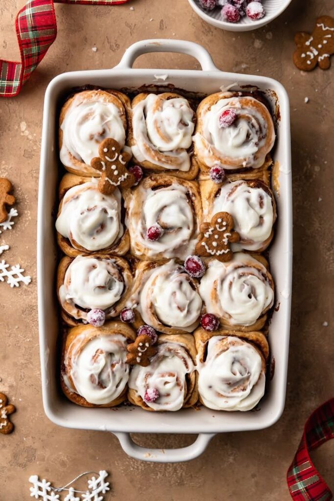 Gingerbread Cinnamon Rolls - Handy Home Festive cinnamon rolls topped with icing, gingerbread cookies, and cranberries in a white baking dish, perfect for the holidays.