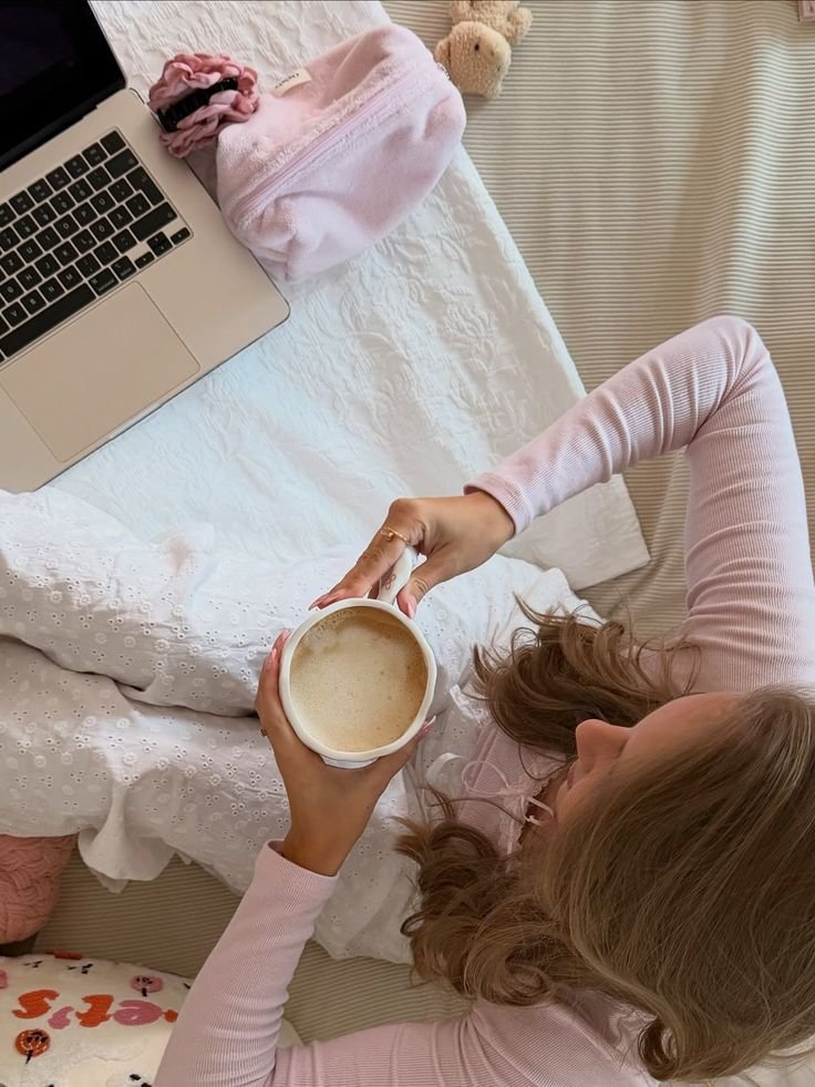 Woman in cozy outfit holding latte, sitting on bed beside laptop and pink pouch. Relaxing morning routine.