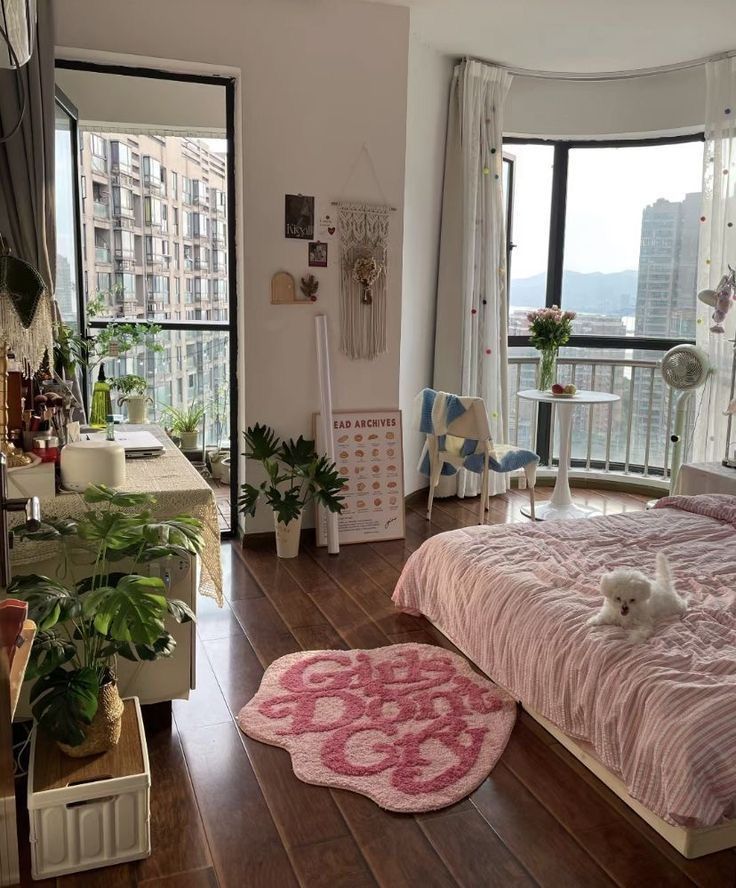 Cozy modern bedroom with pink bedding, plants, a fluffy rug, and large windows overlooking city buildings.