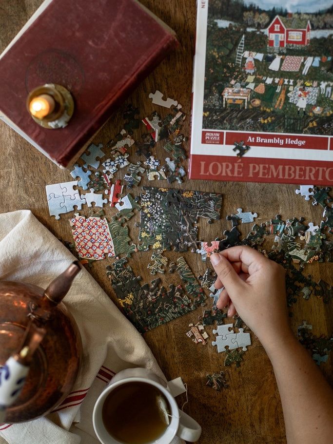 Loré Pemberton Puzzles - Handy Home Hand assembling a jigsaw puzzle with tea and candle on a wooden table. Cozy, relaxing atmosphere.