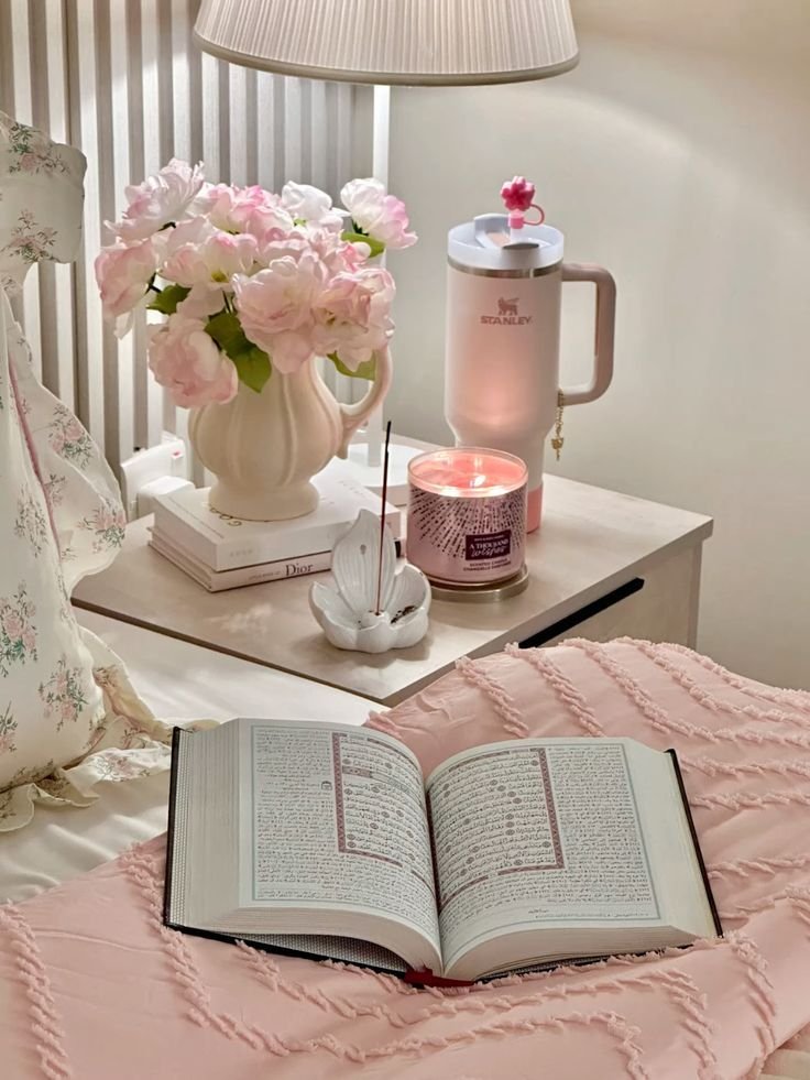 Cozy bedroom scene with open book, pink flowers, a lit candle, and a tumbler on a nightstand, creating a relaxing ambiance.