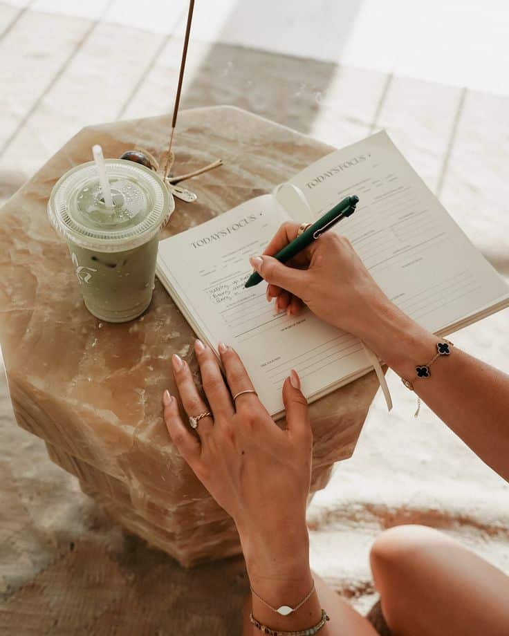 Shes here 🧚🏻 The Flowness Club Journal has - Handy Home Woman journaling with a green pen next to an iced matcha on a marble table, focusing on daily priorities and mindfulness.