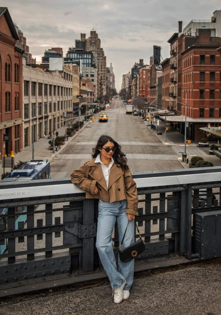 The 10 Best Instagram Spots in New York City _ Anoushka Probyn - Handy Home Stylish woman in sunglasses and brown jacket posing on a city street bridge, with urban skyline and taxis in the background.