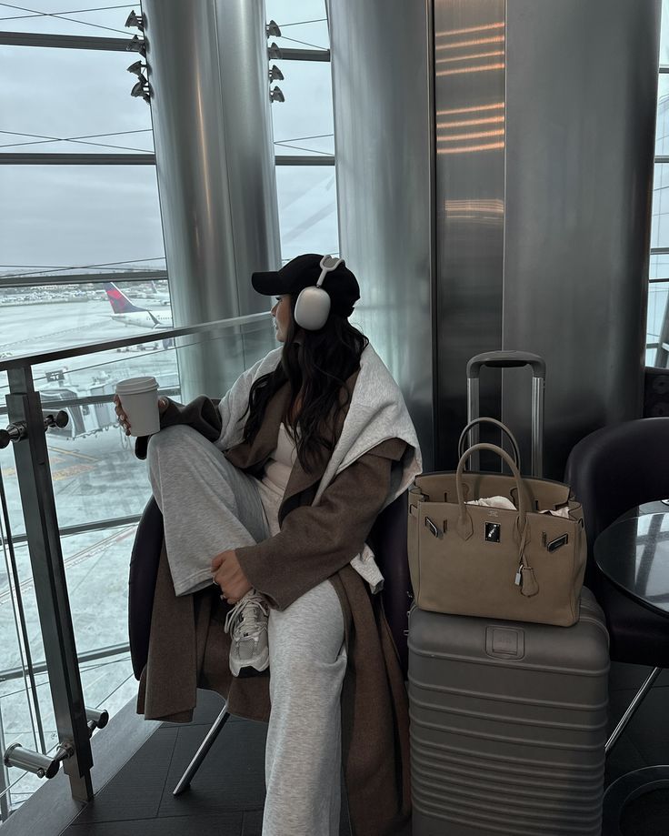 Travel day diaries 🤍 - Handy Home Woman at airport lounge wearing headphones, holding coffee, with luggage and a bag. Airplane in view through the window.