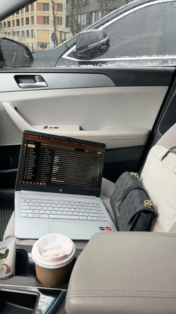 Working 💅🏻 - Handy Home Open laptop on car seat with rainy view, coffee, and purse, creating a cozy mobile workspace.