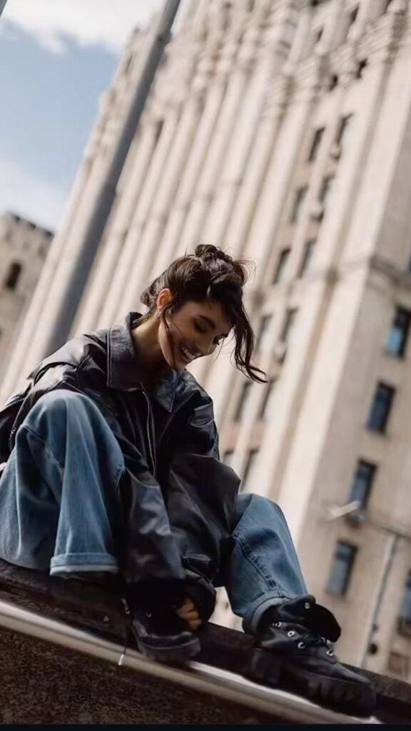 aa2a7e2c-6d1b-4846-b66f-1191b9b4e779 - Handy Home Young woman in oversized denim and leather jacket, smiling on a ledge with an urban building in the background.