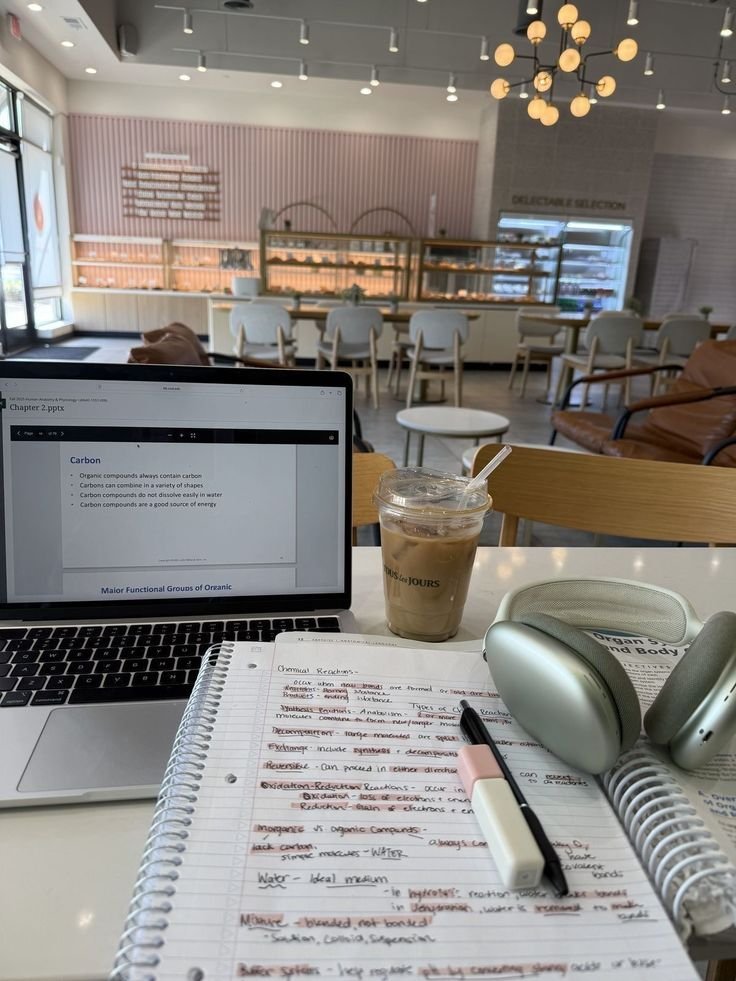 Laptop, iced coffee, notes, and headphones on a cafe table for productive studying. Cozy study spot with natural light and modern decor.