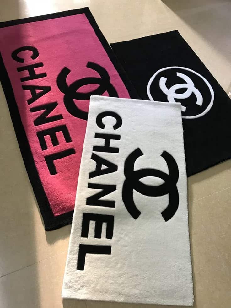 Three Chanel logo rugs in pink, white, and black arranged on a floor, showcasing luxury fashion style and brand identity.