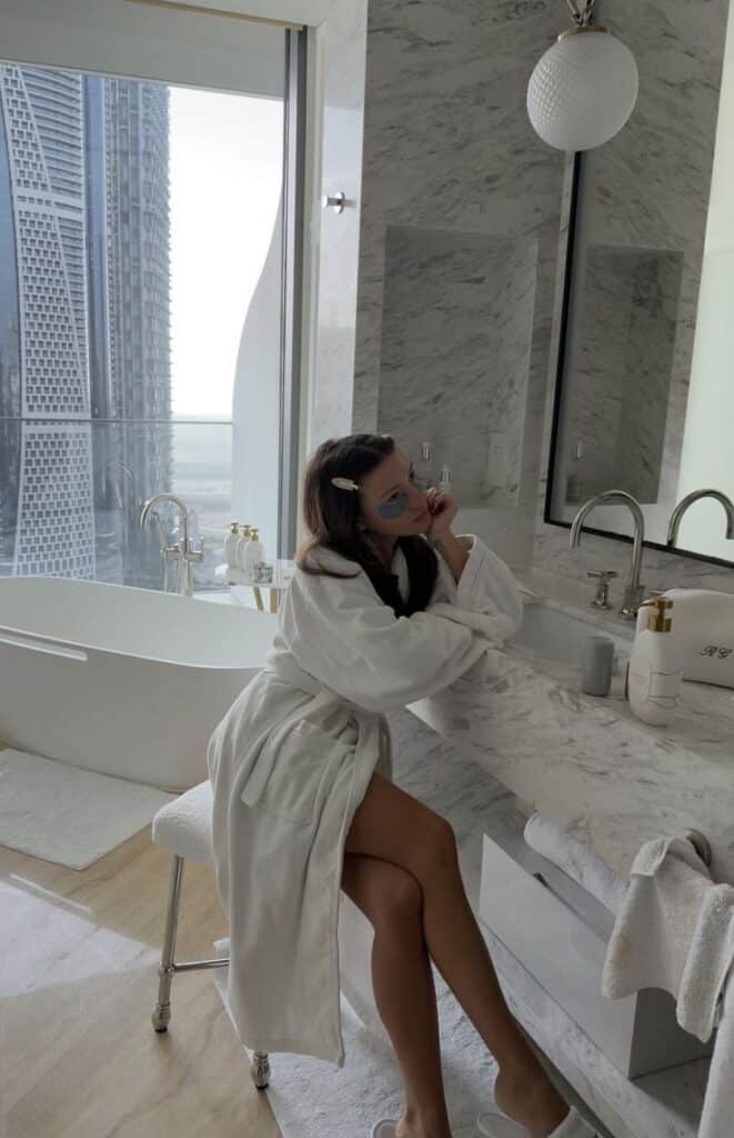 Screenshot - Handy Home A woman in a white robe applying beauty patches in a luxurious marble bathroom with a city view.