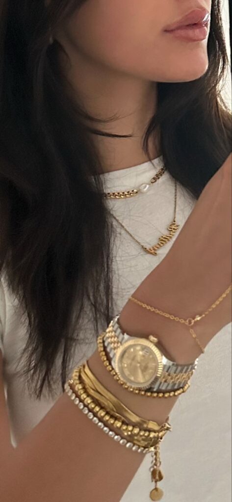 bc443e0d-65ce-4980-b224-ea7174691e0c - Handy Home A woman wearing layered gold necklaces and bracelets, featuring a gold watch, showcases elegant jewelry styling against a white top.