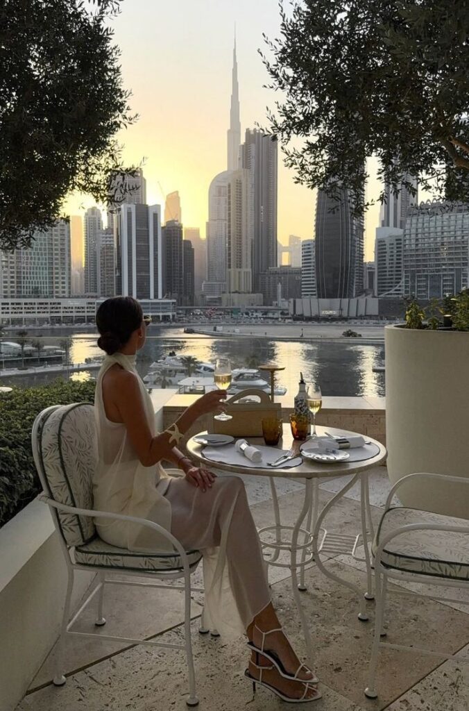 bdce6b4b-df3b-44db-a300-4d71a09d0164 - Handy Home Woman enjoying a sunset dinner with a cityscape view of tall skyscrapers and a tranquil waterfront.
