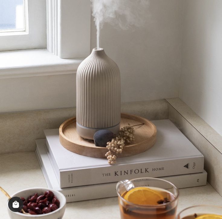 bf5dbdbf-ef75-4a6f-85a9-cc35ebd465a8 - Handy Home Ceramic diffuser emitting mist on a wooden tray with books, by a window. Cozy home decor with a warm beverage and berries nearby.