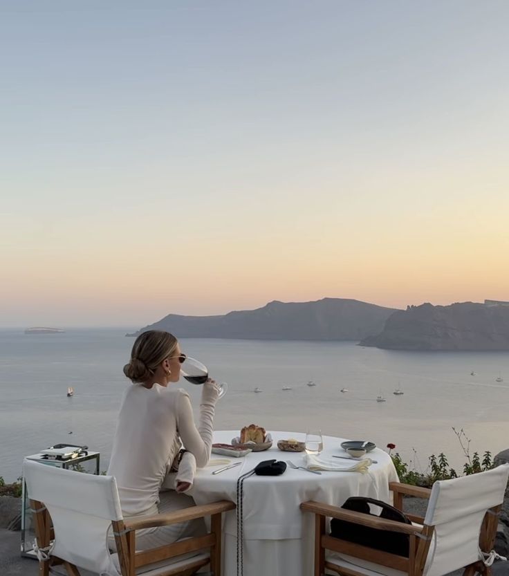 c09e0cad-7bbd-4b31-bc7c-7ea5daad0b6e - Handy Home Woman enjoys sunset view over the ocean while sipping wine at an outdoor dining setup, with coastal hills in the background.