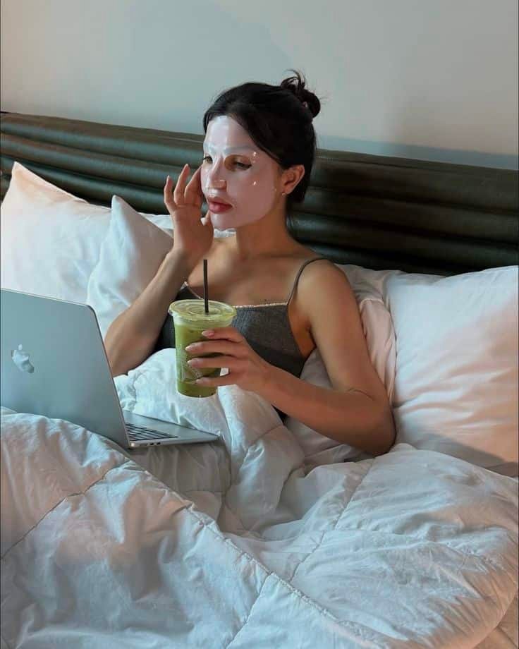 Woman in bed with face mask, drinking green smoothie, working on a laptop, promoting relaxation and self-care while staying connected.