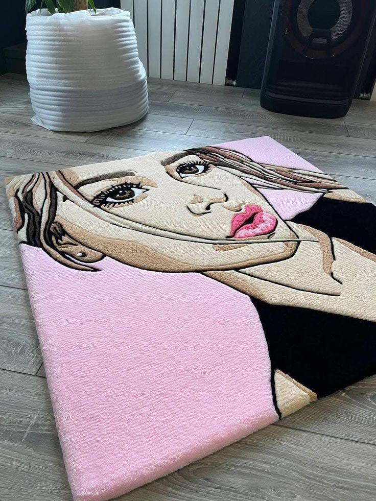 Artistic rug with illustrated female portrait on a pink background, placed on wooden floor near a plant and speaker.