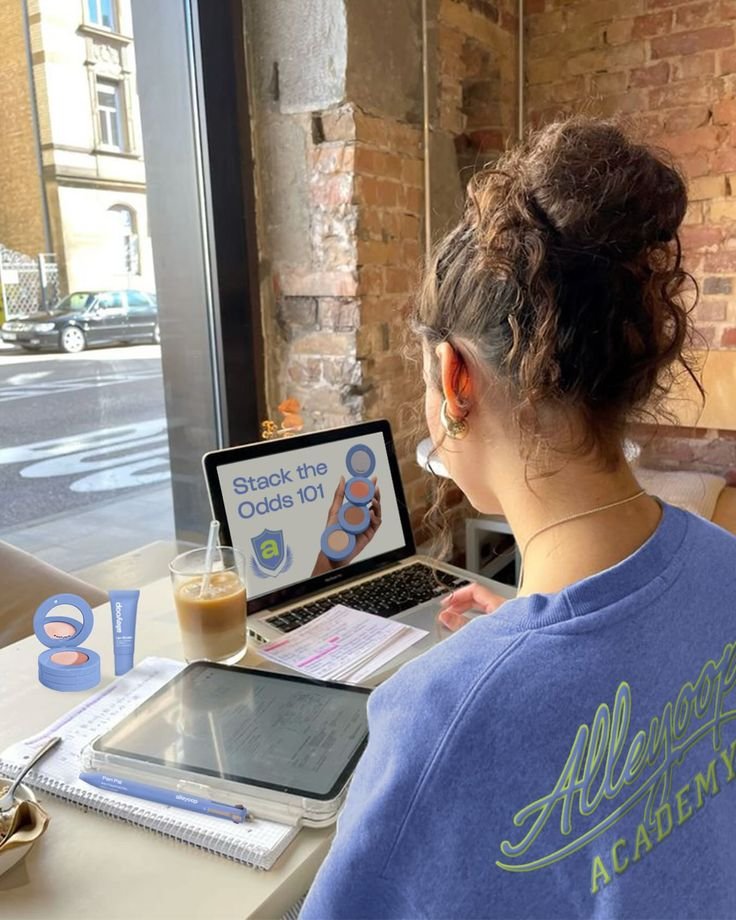 Woman studying with laptop and tablet in a cozy cafe, learning Stack the Odds 101, wearing blue Allegro Academy shirt.