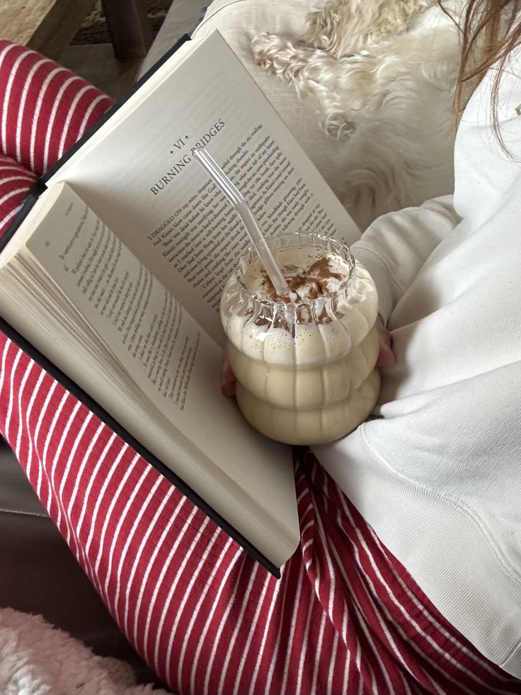 d9db6db9-ba59-45e6-9d05-bd57ee6dea20 - Handy Home Cozy reading with a book, latte, and a fluffy dog on a sofa. Red striped pants add a touch of warmth to the relaxing scene.