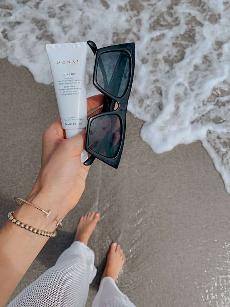df00733f-312b-4548-8509-9319a5b67cd4 - Handy Home Hand holding sunscreen and sunglasses on a sandy beach, with gentle waves in the background. Perfect summer essentials.