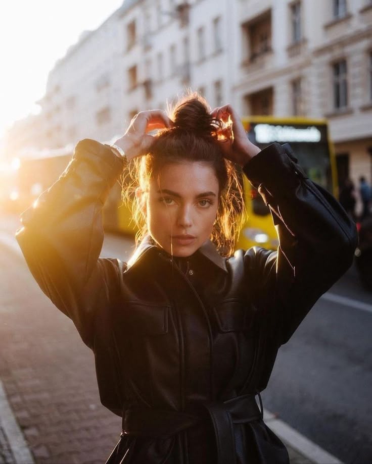 e110fc35-ab1f-4870-8377-30589c5b4407 - Handy Home Woman in leather jacket adjusts hair in city street at sunset, with yellow buses and buildings in the background.