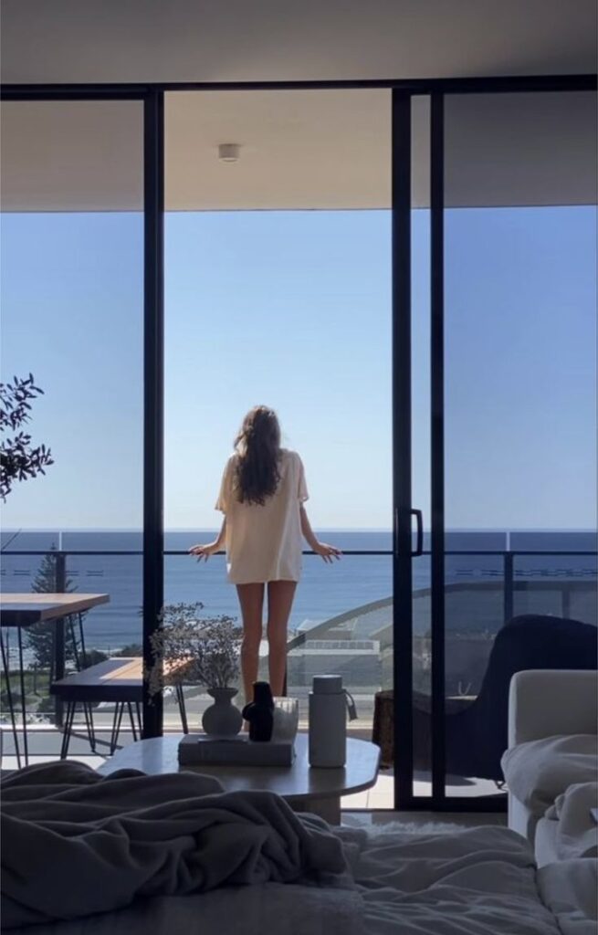 Screenshot - Handy Home Woman in a t-shirt enjoying a sunny ocean view from a modern apartment balcony, captured through large glass doors.