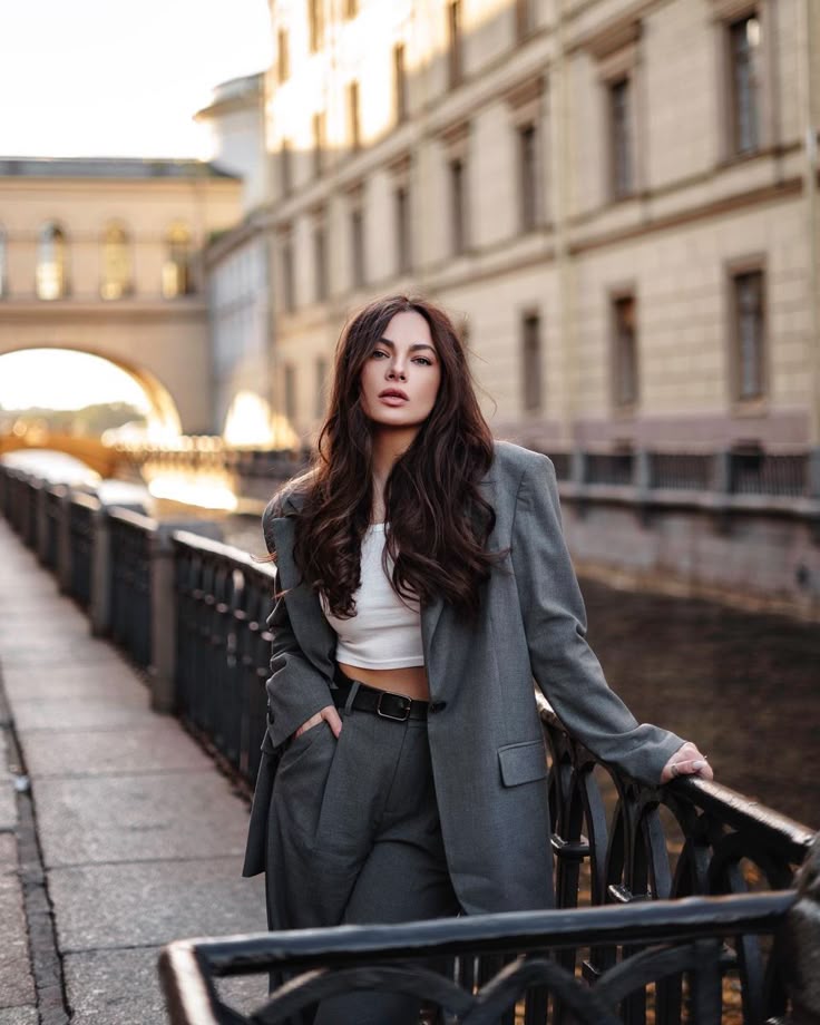 e9b7cc53-bccb-47fd-8659-dc6c94b88ff6 - Handy Home Woman in stylish gray suit poses confidently on city bridge walkway, with historic architecture in the background.