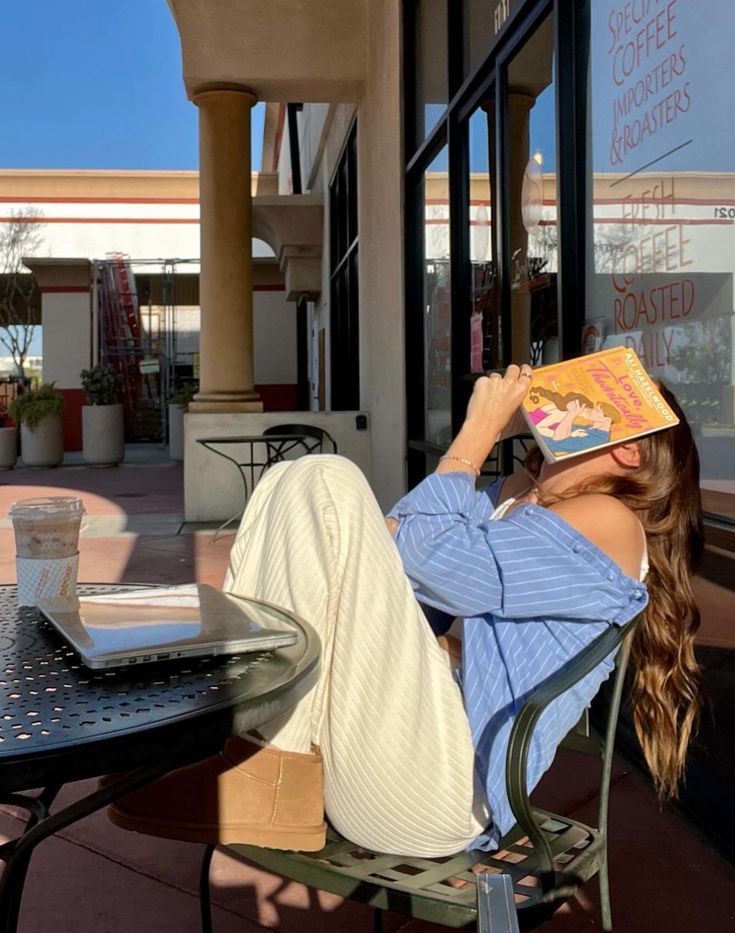 ee0c92a4-312b-4fd2-89c0-1c614ded5be8 - Handy Home Woman in a blue shirt relaxing outside a cafe with a book, next to a laptop and coffee, enjoying a sunny day.