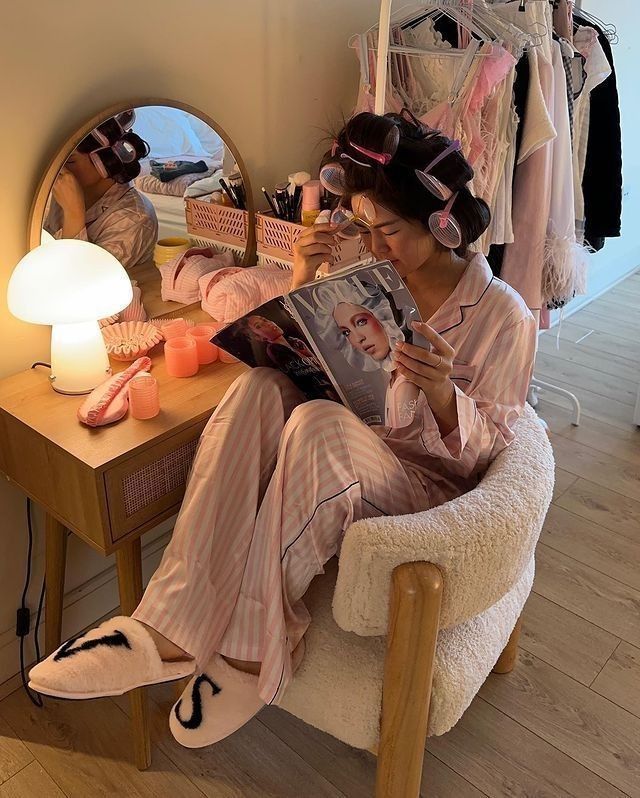 f2b1313b-b064-4ba5-a0af-d48d0b3b1650 - Handy Home Woman in pajamas with hair rollers reads a fashion magazine at a cozy vanity, surrounded by makeup and a soft lamp.