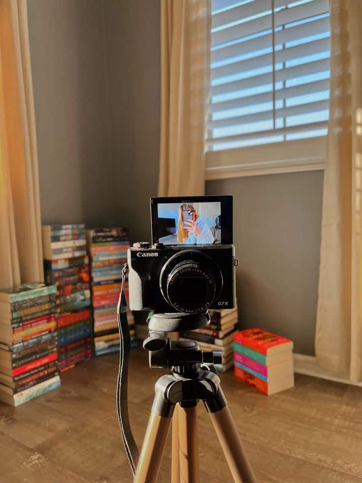 ff7b425c-2cbb-48cc-ae3e-1290dba707a6 - Handy Home Camera on tripod with selfie on screen in cozy room, stacks of books in background by the window. Perfect for home photography.