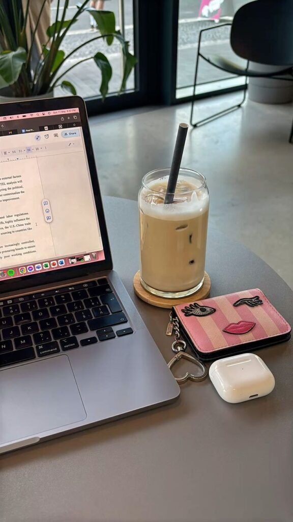 Laptop and iced coffee on a cafe table with a quirky pink wallet and AirPods, perfect for casual workday vibes.