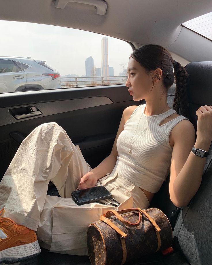 h_yeli - Handy Home Woman in car gazing outside, wearing white top and pants, with a designer bag and smartphone on her lap. Urban skyline in background.