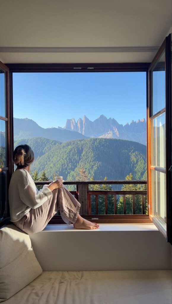 italy - Handy Home Woman sitting by an open window, enjoying a mountain view with a cup of tea at sunrise, serene and relaxing nature setting.