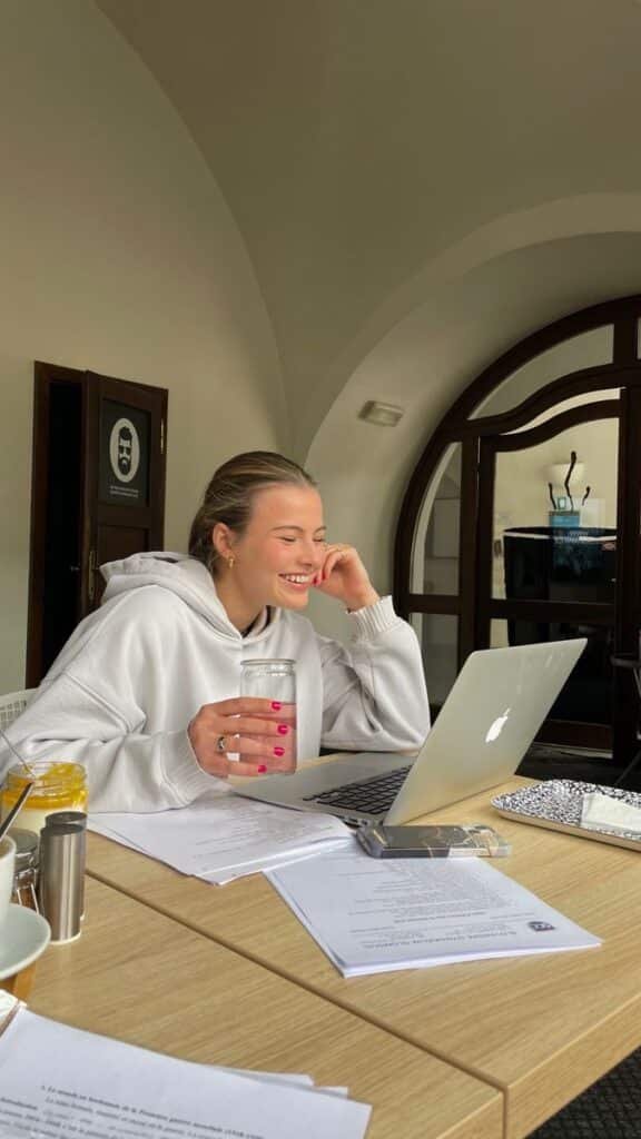 Smiling woman in a white hoodie works on a laptop, holding a drink at a cozy cafe table.
