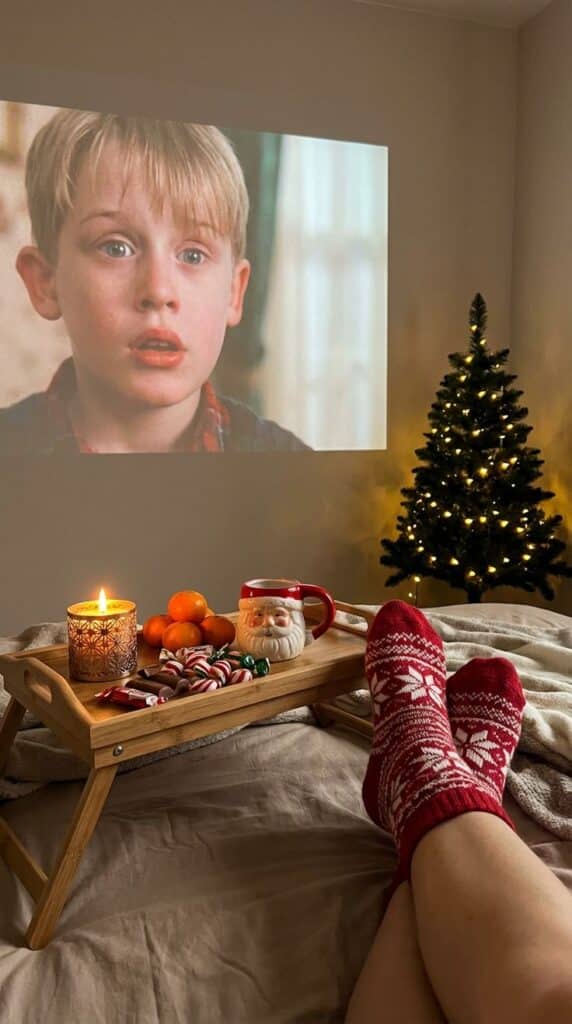 zdgnzdbs - Handy Home Cozy Christmas scene: movie projected on wall, festive socks, decorated tree, Santa mug, candlelight, and snacks on a wooden tray.