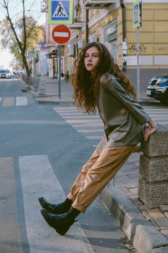 Young woman with curly hair leans on a street post at an urban crosswalk, exuding casual style in a relaxed outfit.