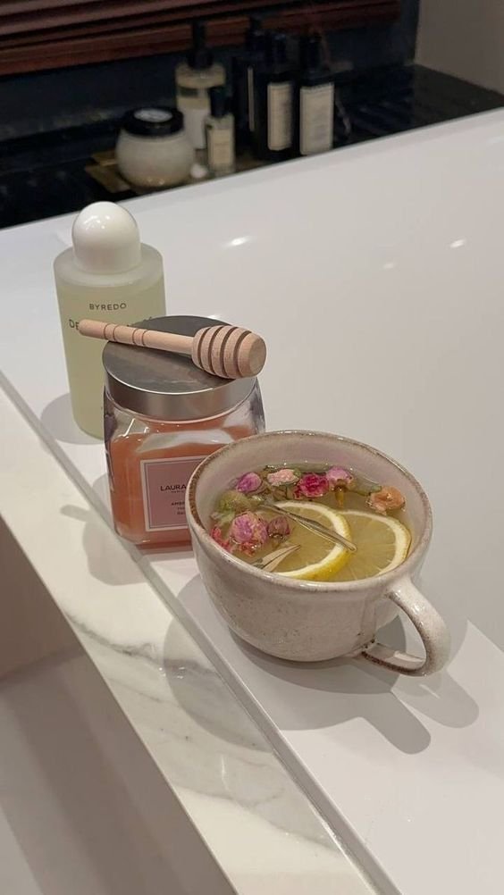 Cozy spa setting with herbal tea, honey, candle, and luxury skincare on a marble bathtub ledge for relaxation and aromatherapy.