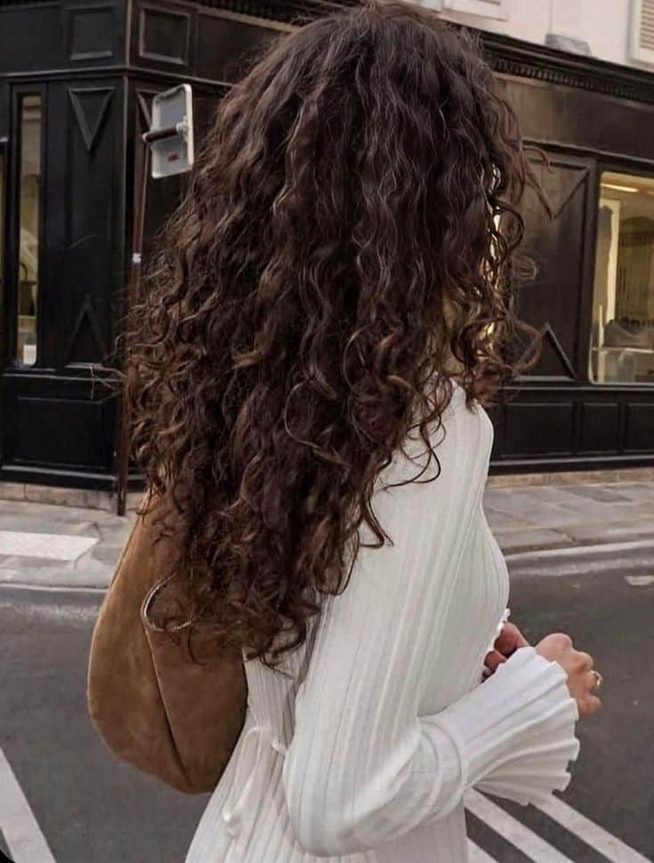 0a9c6563-9309-49a3-aad5-20cc0055e4b5 Woman with long curly hair walks on city street in a white dress, carrying a brown suede bag. Urban fashion style. - Handy Home Woman with long curly hair walks on city street in a white dress, carrying a brown suede bag. Urban fashion style.