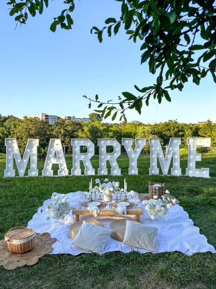Romantic outdoor marriage proposal setup with Marry Me lights, a picnic blanket, flowers, and candles under a tree.