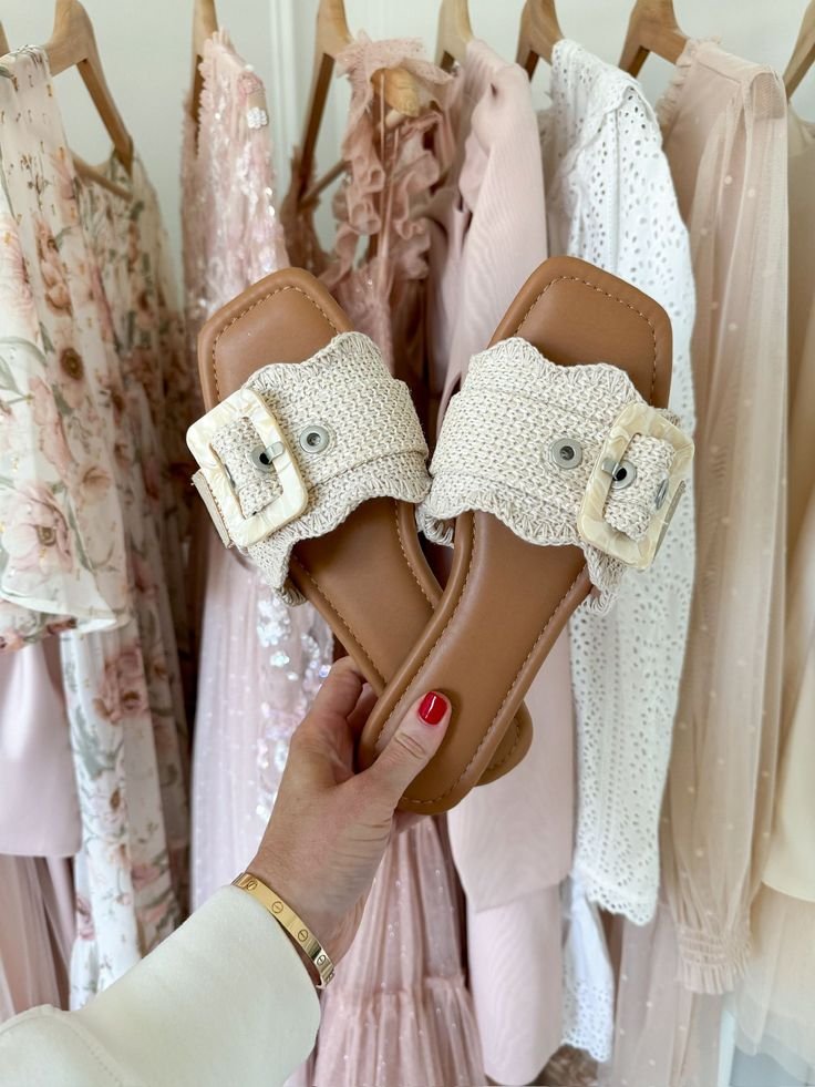 Hand holds stylish woven sandals with beige buckle, in front of pastel dresses on hangers. Fashion and summer style concept.