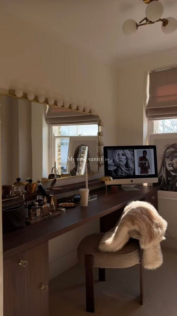 Stylish vanity setup with a large mirror, computer, makeup, and a cozy fur-draped chair in a well-lit room.