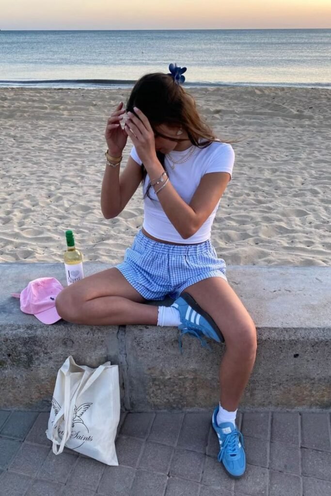 Young woman sitting by the beach at sunset, wearing casual summer clothing, with a bottle of wine and a tote bag beside her.