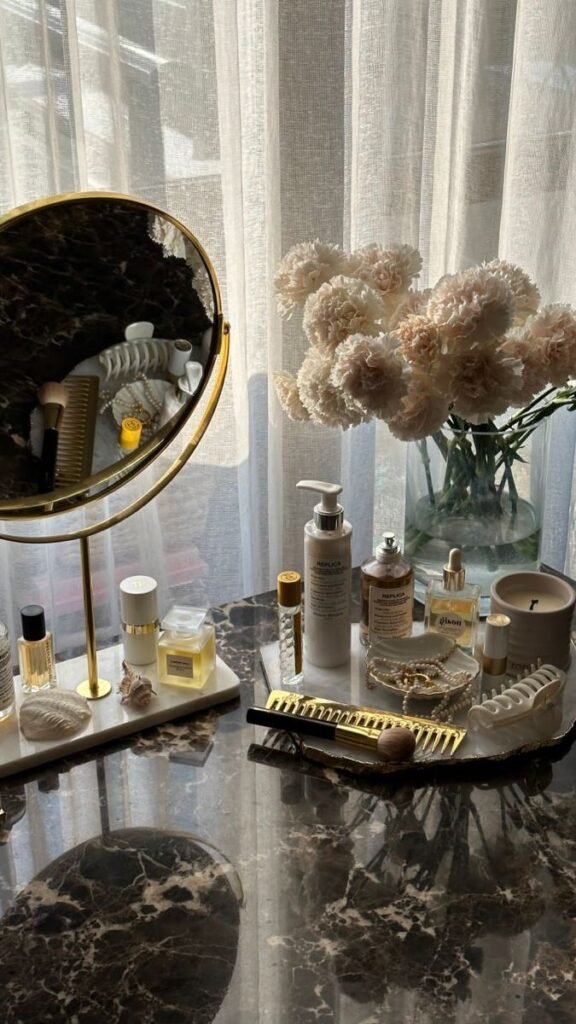 Elegant vanity setup with beauty products, gold accents, and a vase of flowers on a marble surface by a window with sheer curtains.