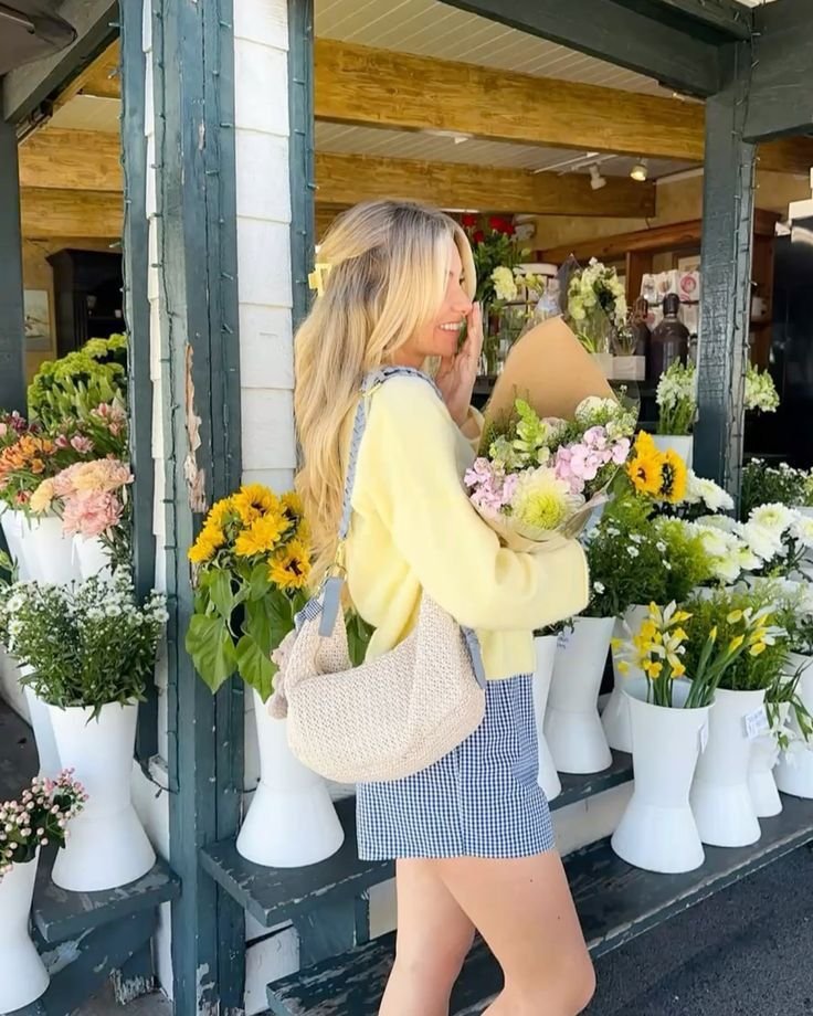 A woman in a yellow sweater holding a bouquet of flowers, standing outside a vibrant flower shop with white vases of blooms.
