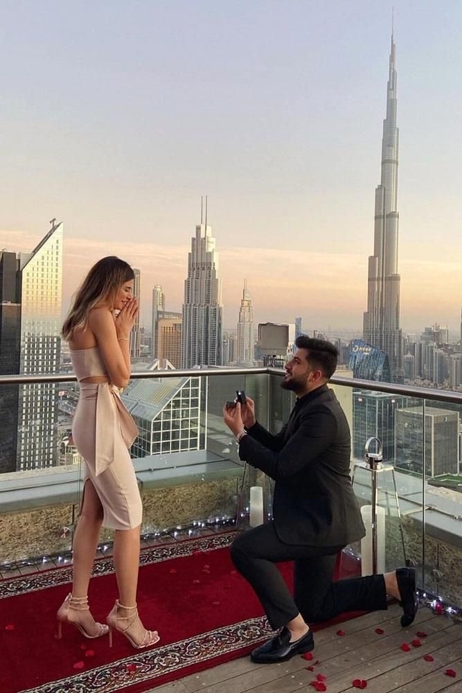 Man proposing on rooftop with a ring, woman surprised, Burj Khalifa skyline background, romantic cityscape moment.
