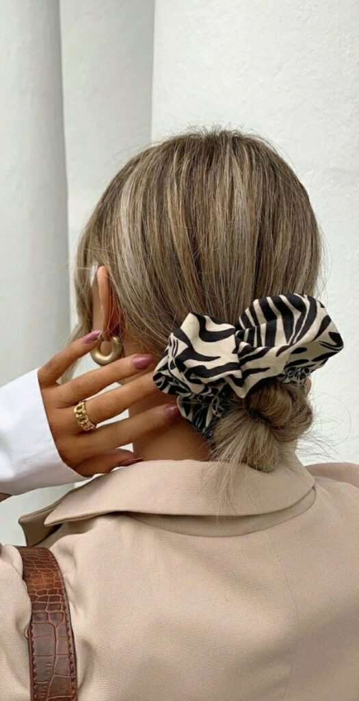 29c7190a-0bb0-432a-b182-5b897a8fe748 Woman with zebra-print scrunchie holding hair in low bun, wearing gold rings, gold hoop earring, and beige blazer. - Handy Home Woman with zebra-print scrunchie holding hair in low bun, wearing gold rings, gold hoop earring, and beige blazer.