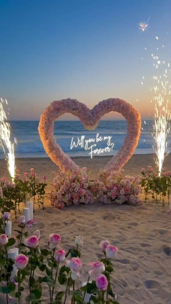 Romantic beach proposal setup with pink roses and floral heart arch at sunset, glowing Will you be my forever? sign.
