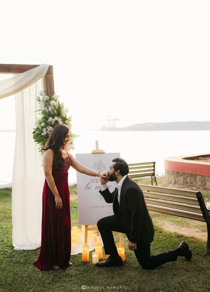 Man proposing to woman in elegant outdoor setting by the lake, surrounded by candles and flowers, romantic gesture.