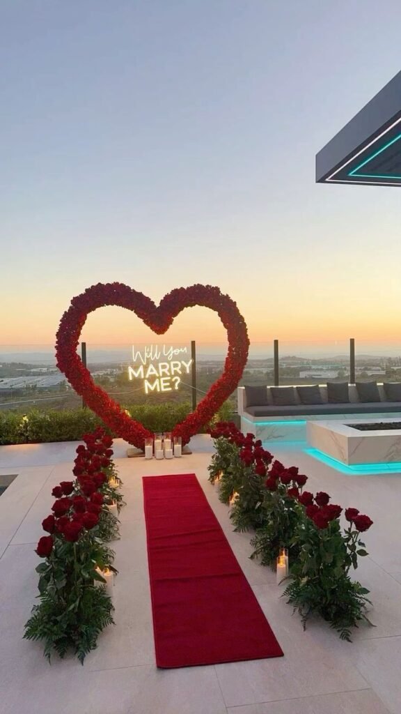 Romantic sunset proposal with a heart-shaped rose arch and Will You Marry Me? sign, red carpet, and roses lining the walkway.