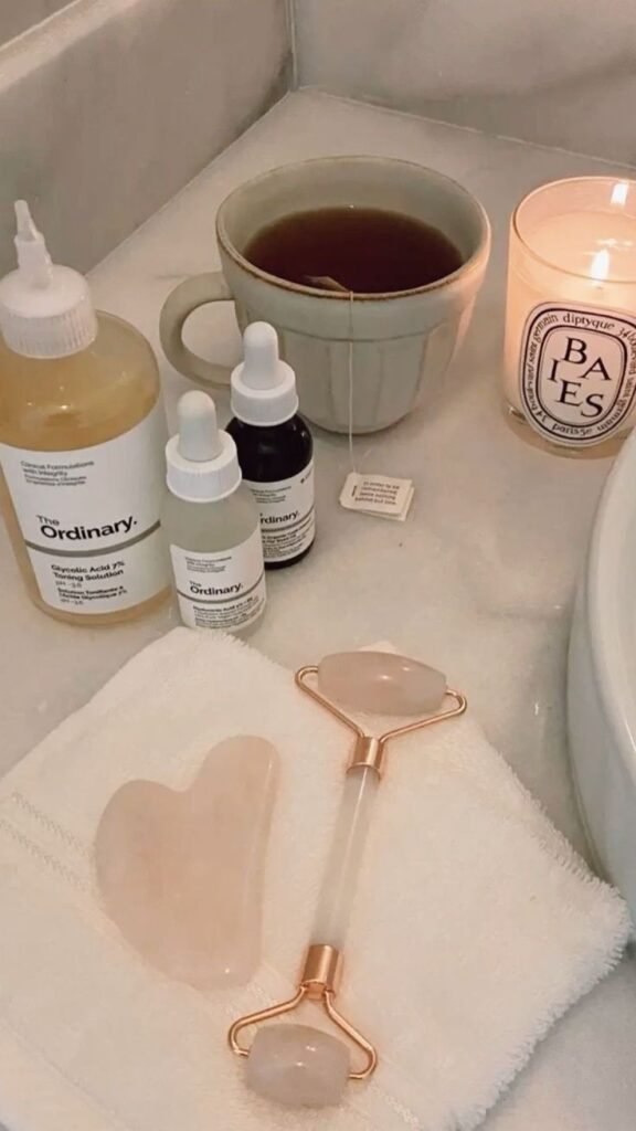 Skincare setup with The Ordinary products, candle, jade roller, gua sha, tea, on white towel. Relaxing self-care routine in bathroom.