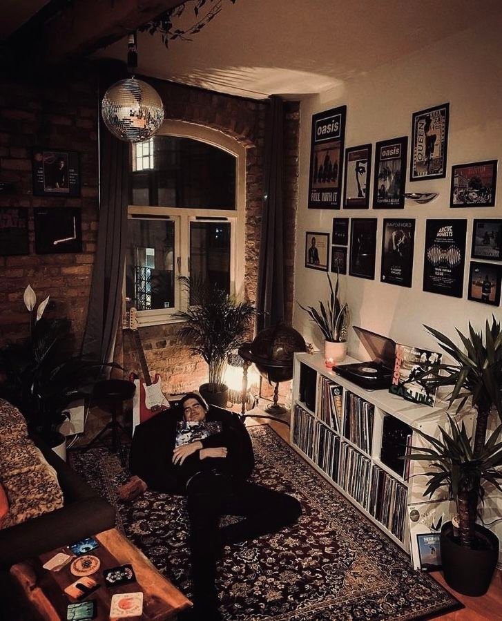 Cozy room with records, guitar, disco ball, and ambient lighting. Person relaxing on a patterned rug by a music-themed wall.